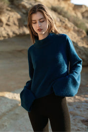 Woman wearing a petrol blue cashmere sweater by Nuage Cashmere on the beach, showing the front with wide sleeves and a high neck