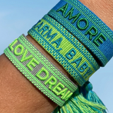 Close-up of adjustable azurro blue woven friendship bracelet with green embroidered "AMORE" lettering, matching green borders, and sliding knot tassel cords.