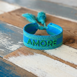 Close-up of adjustable azurro blue woven friendship bracelet with green embroidered "AMORE" lettering, matching green borders, and sliding knot tassel cords.