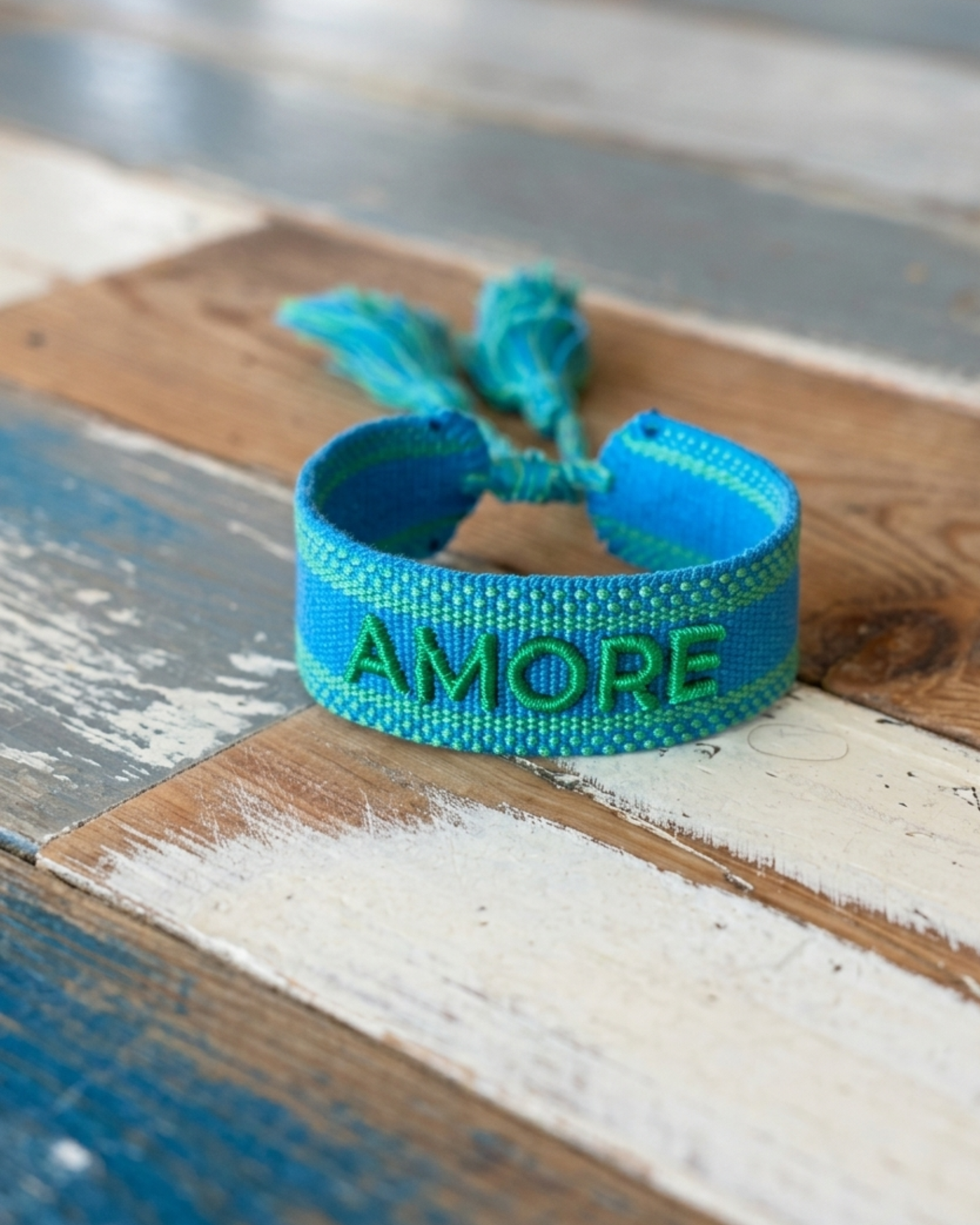 Close-up of adjustable azurro blue woven friendship bracelet with green embroidered "AMORE" lettering, matching green borders, and sliding knot tassel cords.