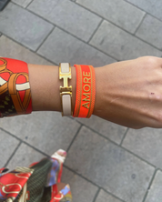 Close-up of adjustable neon orange woven friendship bracelet with green border and gold embroidered "AMORE" lettering and sliding knot cords.