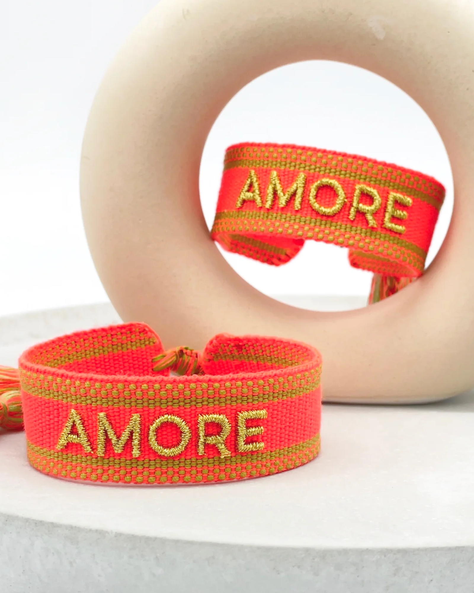 Close-up of adjustable neon orange woven friendship bracelet with green border and gold embroidered "AMORE" lettering and sliding knot cords.