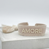 Close-up of adjustable soft taupe woven bracelet with embroidered “AMORE” lettering and sliding knot tassel cords.