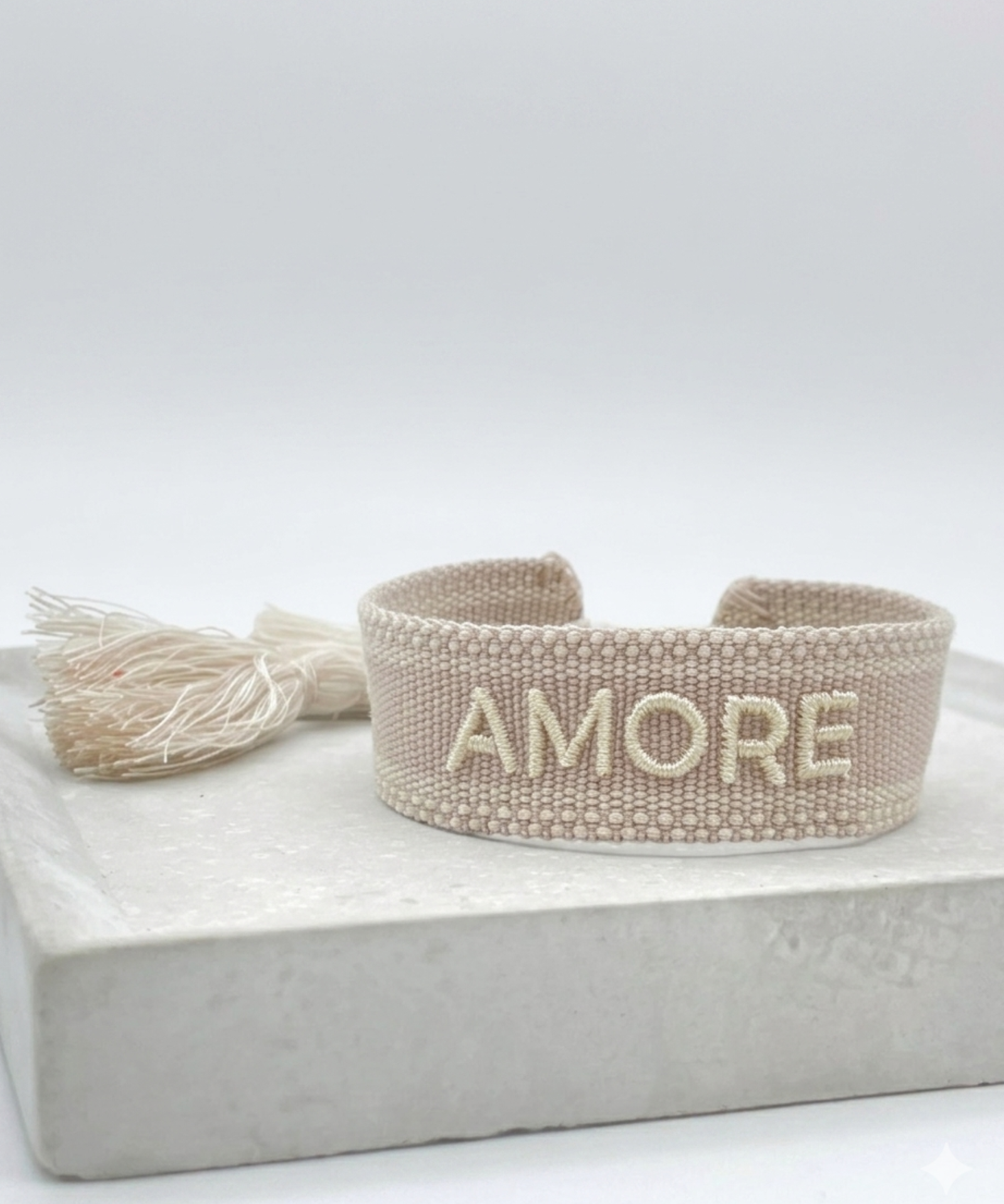 Close-up of adjustable soft taupe woven bracelet with embroidered “AMORE” lettering and sliding knot tassel cords.