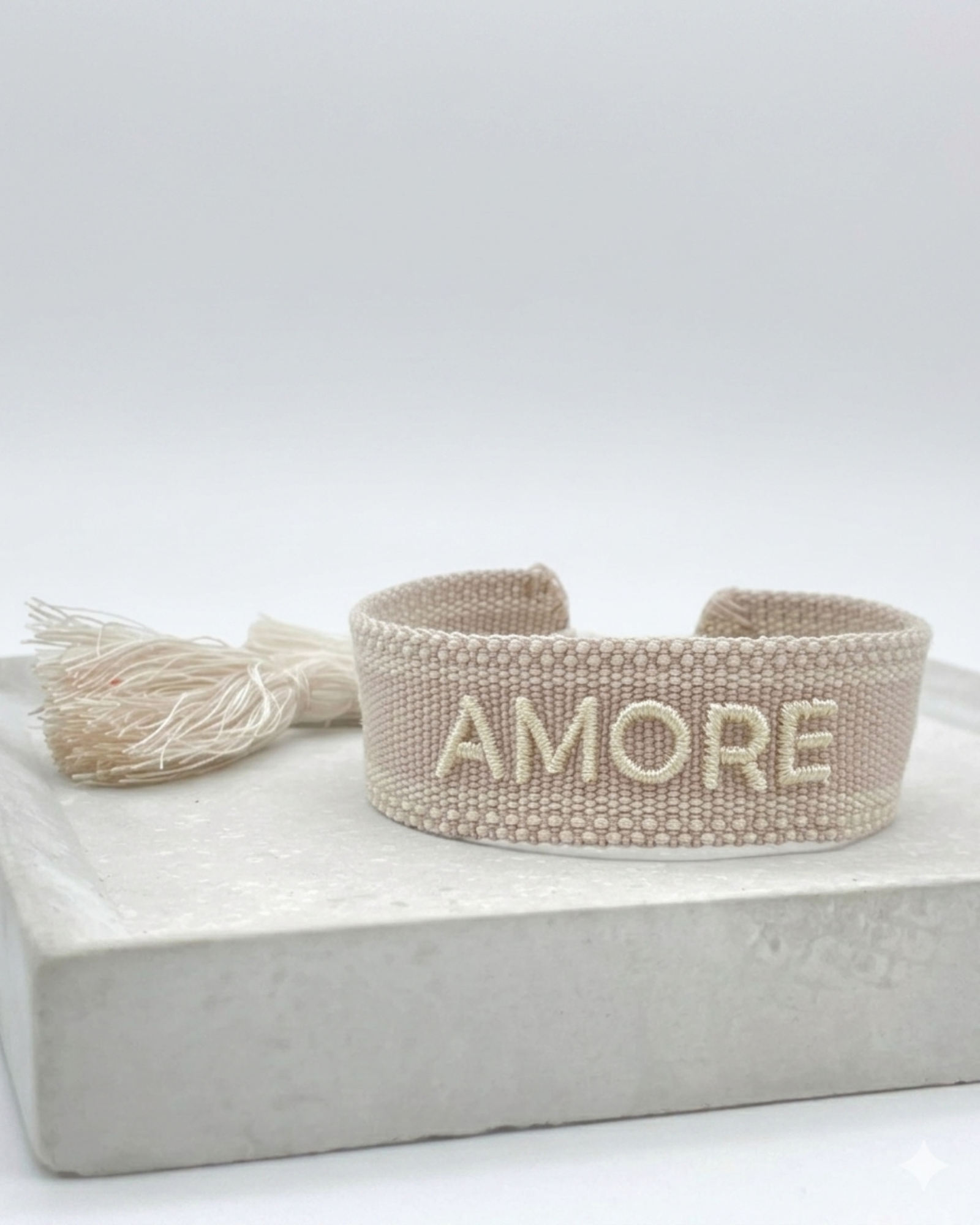 Close-up of adjustable soft taupe woven bracelet with embroidered “AMORE” lettering and sliding knot tassel cords.