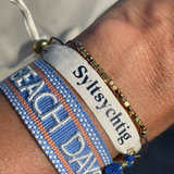 Close-up of adjustable blue woven friendship bracelet with off-white and orange border and embroidered "BEACH DAYS" lettering with sliding knot cords.