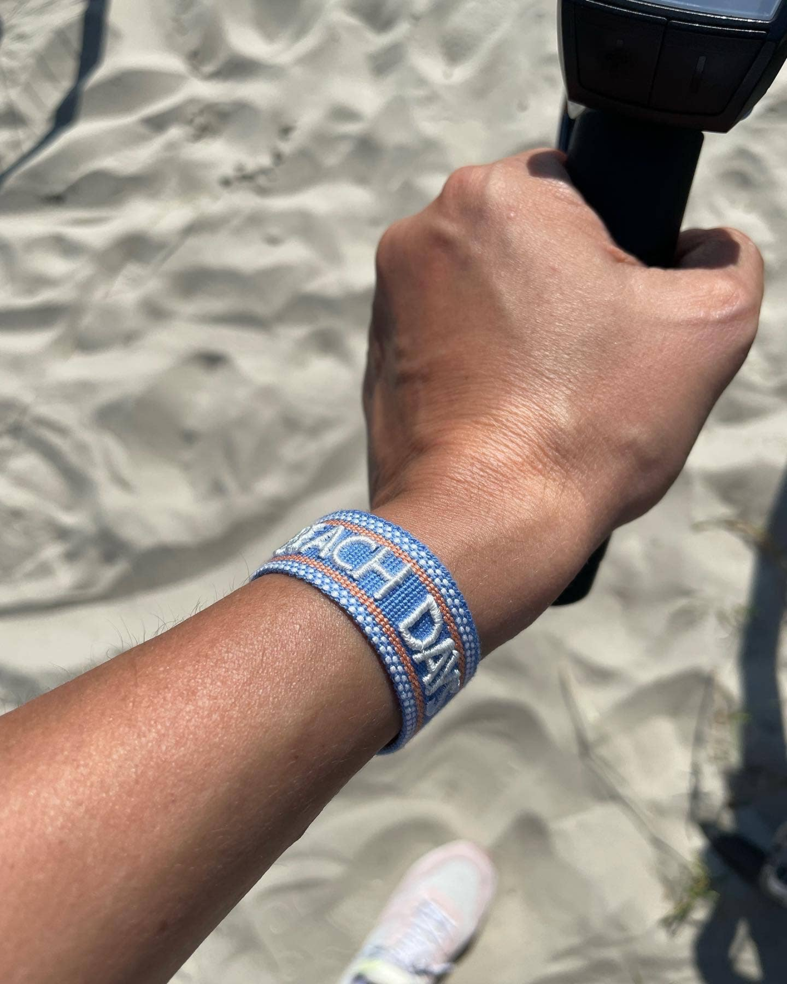 Close-up of adjustable blue woven friendship bracelet with off-white and orange border and embroidered "BEACH DAYS" lettering with sliding knot cords.