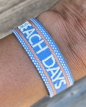 Close-up of adjustable blue woven friendship bracelet with off-white and orange border and embroidered "BEACH DAYS" lettering with sliding knot cords.