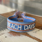 Close-up of adjustable blue woven friendship bracelet with off-white and orange border and embroidered "BEACH DAYS" lettering with sliding knot cords.