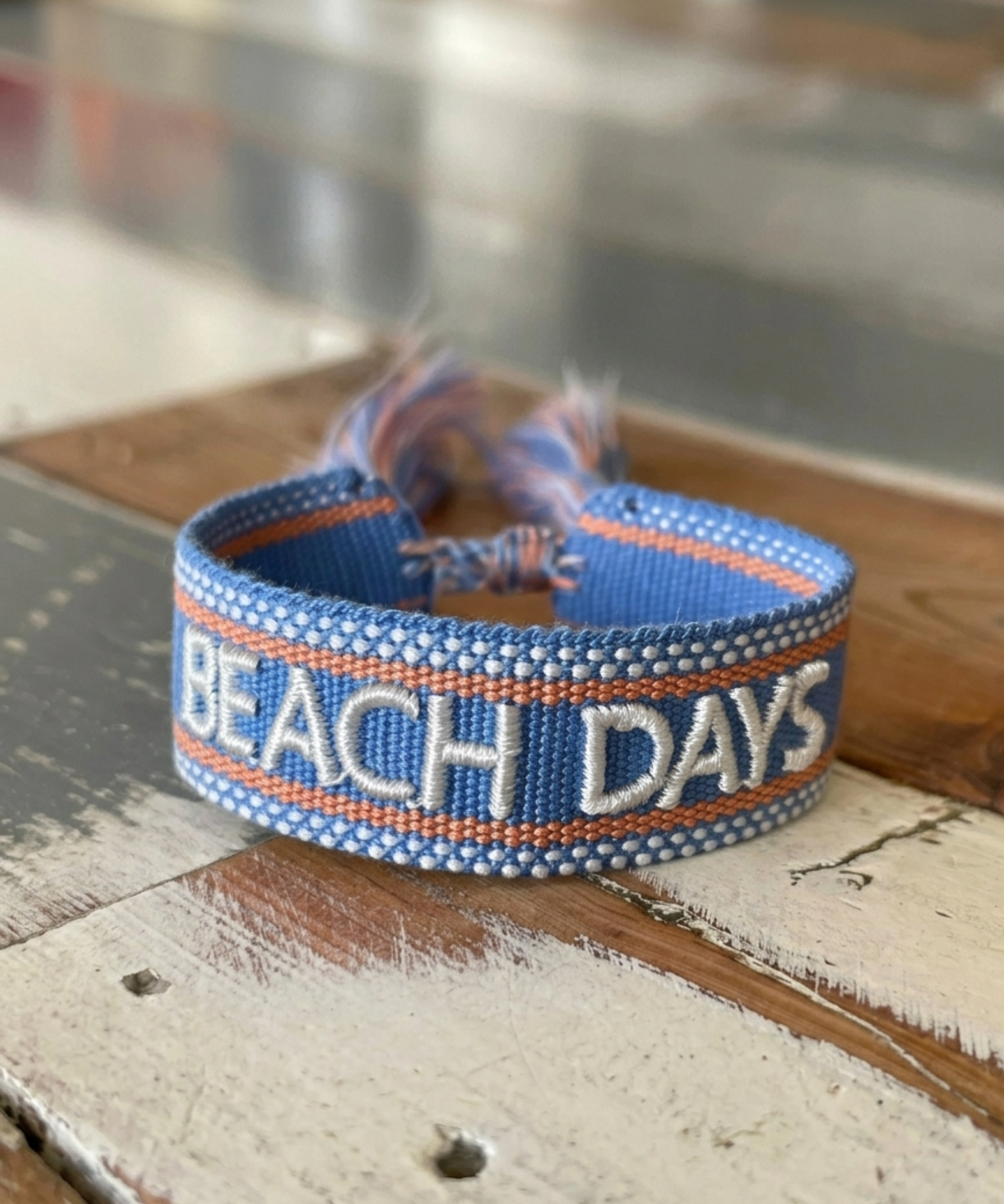 Close-up of adjustable blue woven friendship bracelet with off-white and orange border and embroidered "BEACH DAYS" lettering with sliding knot cords.