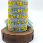 Close-up of adjustable light blue woven friendship bracelet with yellow embroidered "BELIEVE IN MAGIC" text and off-white border with sliding knot cords.