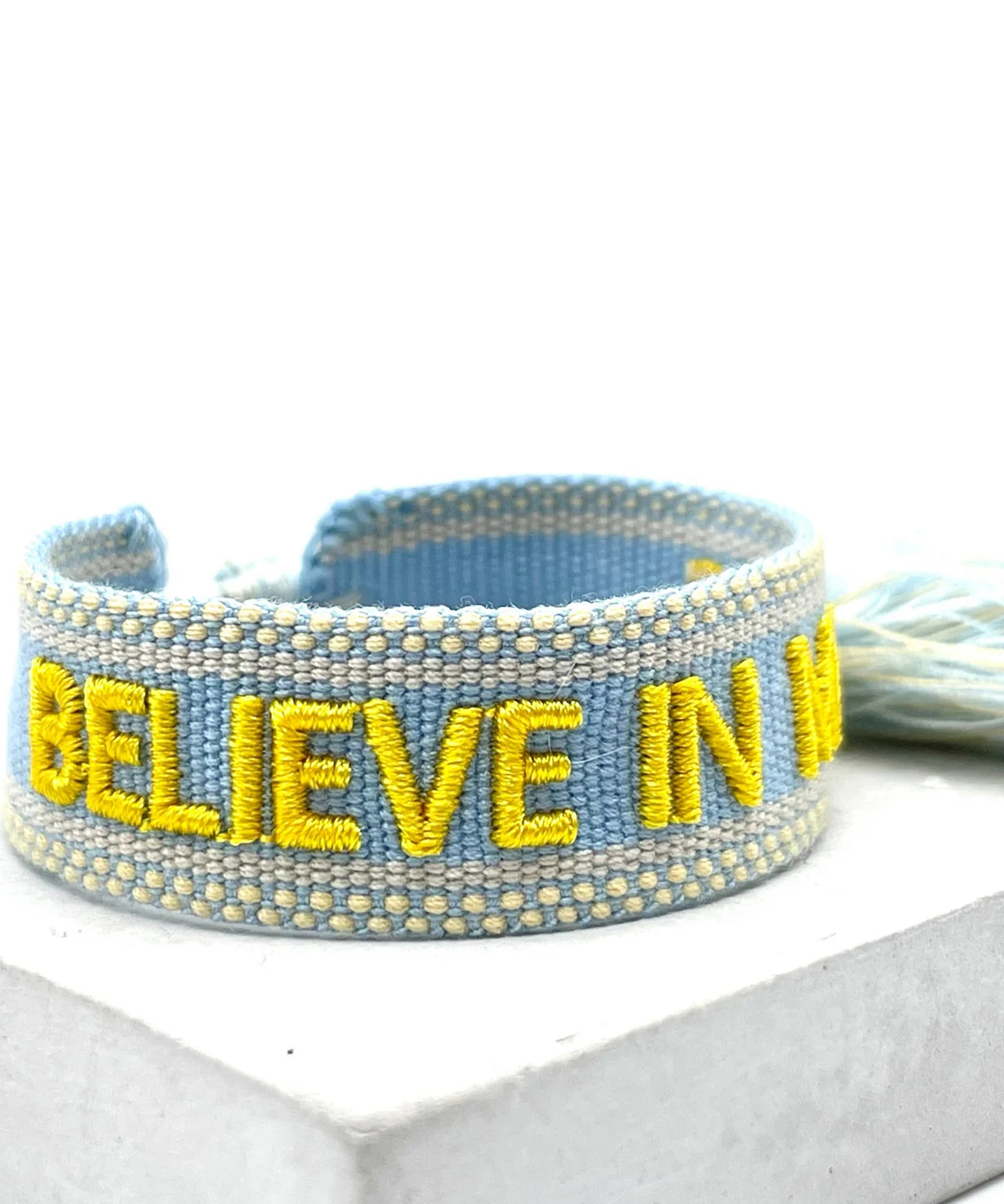 Close-up of adjustable light blue woven friendship bracelet with yellow embroidered "BELIEVE IN MAGIC" text and off-white border with sliding knot cords.