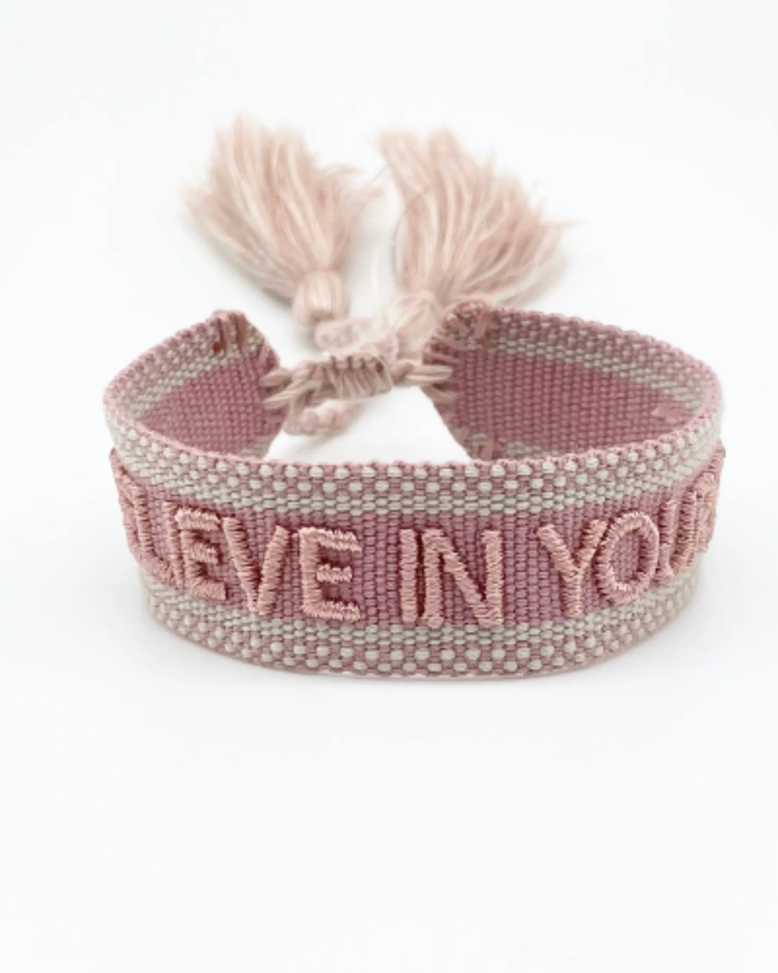 Close-up of adjustable dusty rose woven bracelet with embroidered “BELIEVE IN YOURSELF” lettering and sliding knot tassel cords.