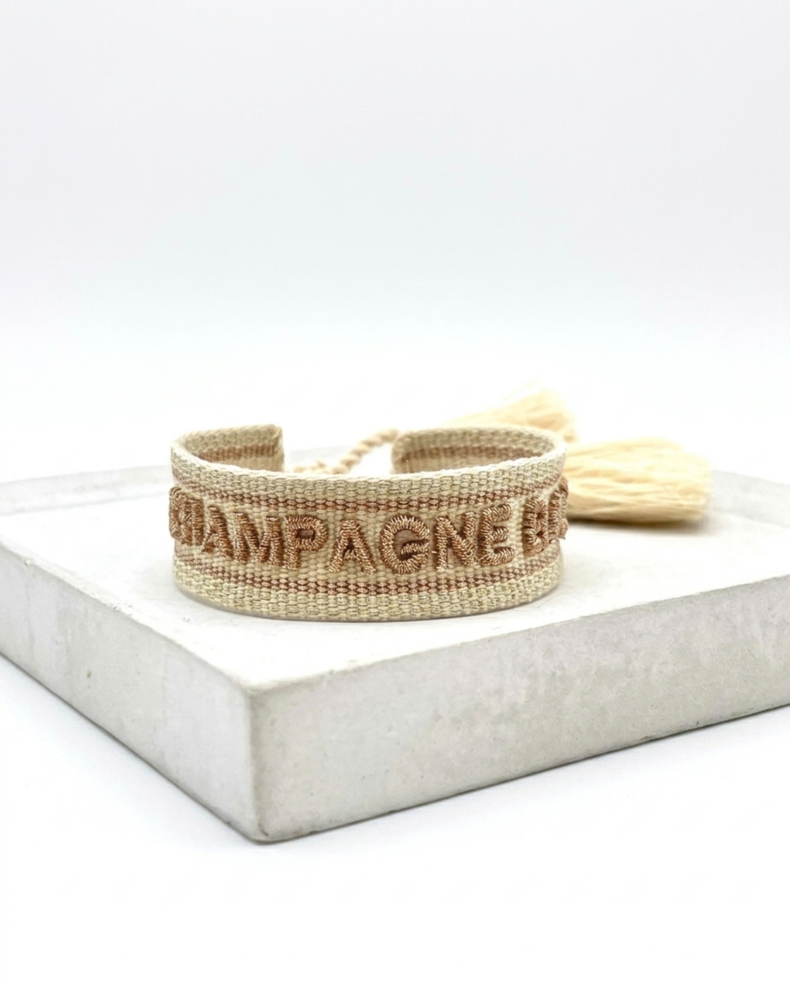 Close-up of adjustable beige woven bracelet with glitter taupe embroidered “CHAMPAGNE BABY” lettering, glitter taupe gold border, and sliding knot tassel cords.
