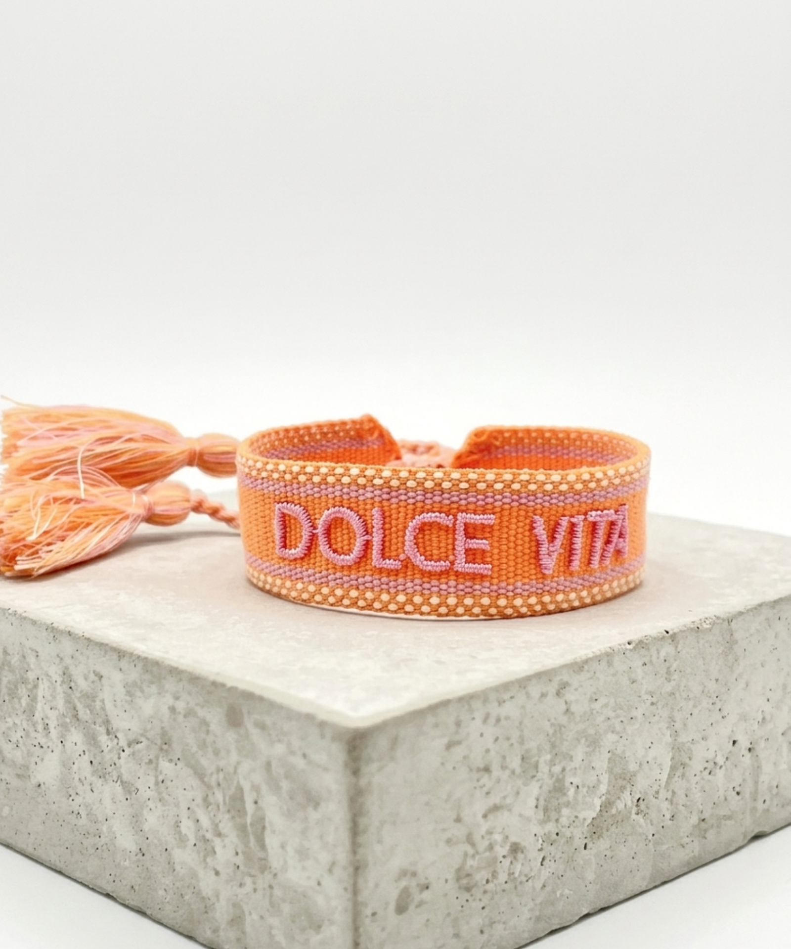 Close-up of adjustable orange woven bracelet with embroidered “DOLCE VITA” lettering and sliding knot tassel cords.