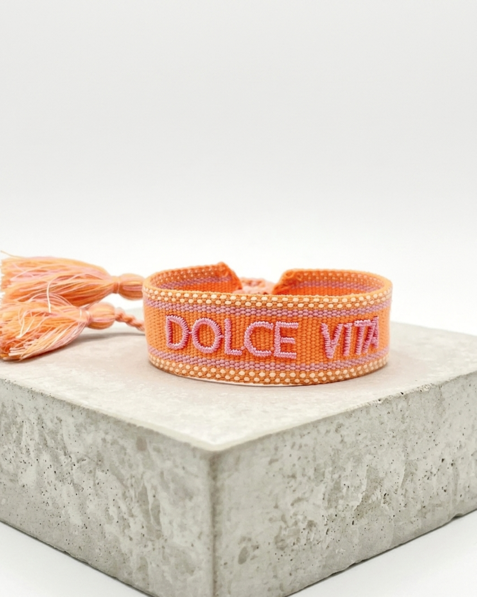 Close-up of adjustable orange woven bracelet with embroidered “DOLCE VITA” lettering and sliding knot tassel cords.