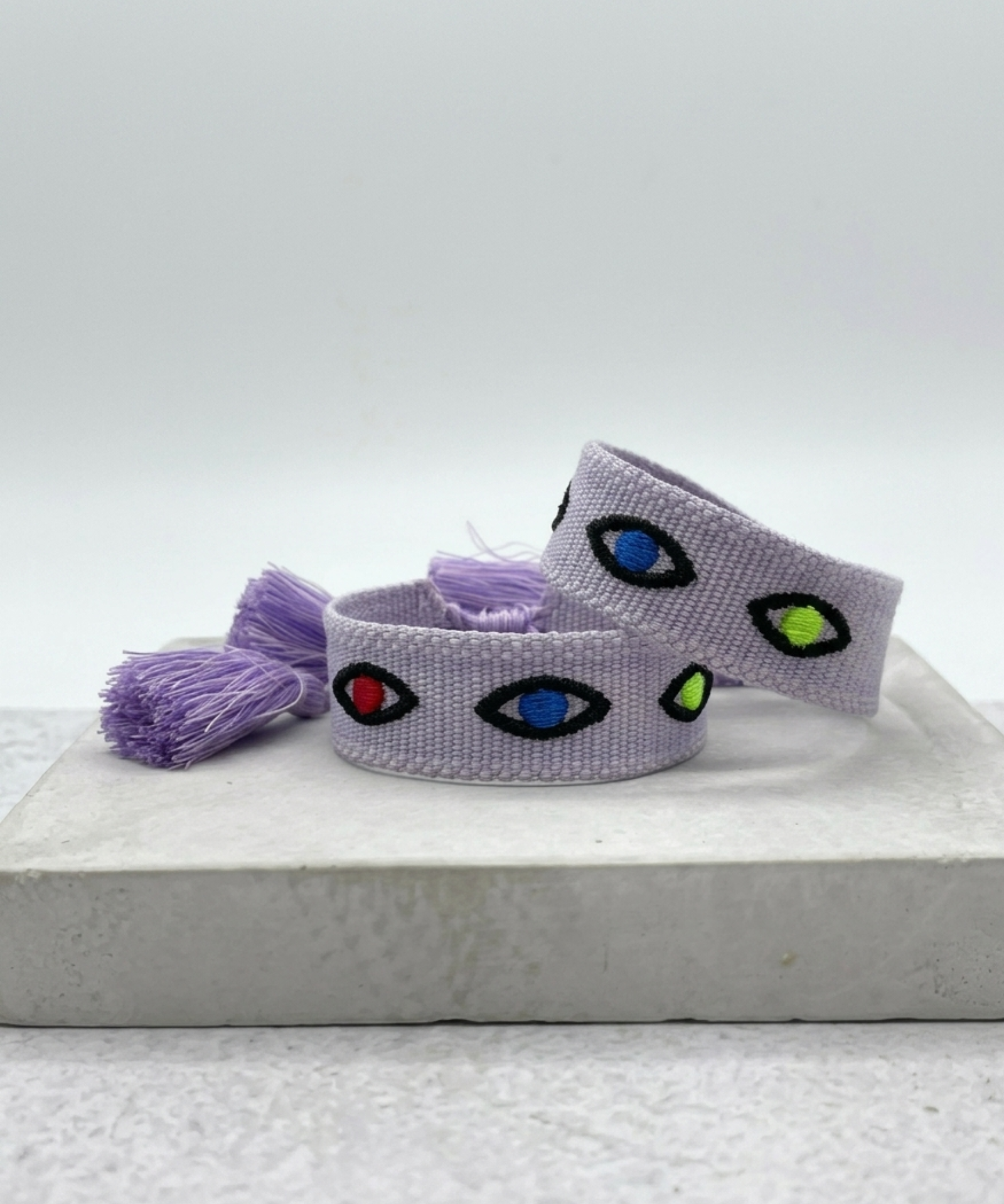 Close-up of adjustable lavender woven bracelet featuring embroidered evil eye talisman design with sliding knot tassel cords.