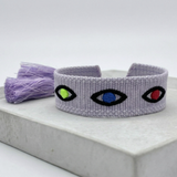 Close-up of adjustable lavender woven bracelet featuring embroidered evil eye talisman design with sliding knot tassel cords.