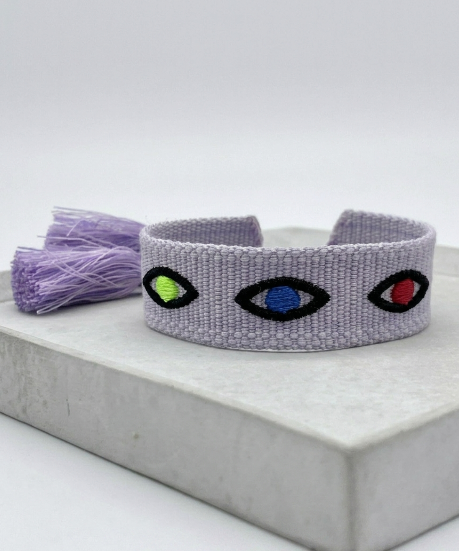 Close-up of adjustable lavender woven bracelet featuring embroidered evil eye talisman design with sliding knot tassel cords.