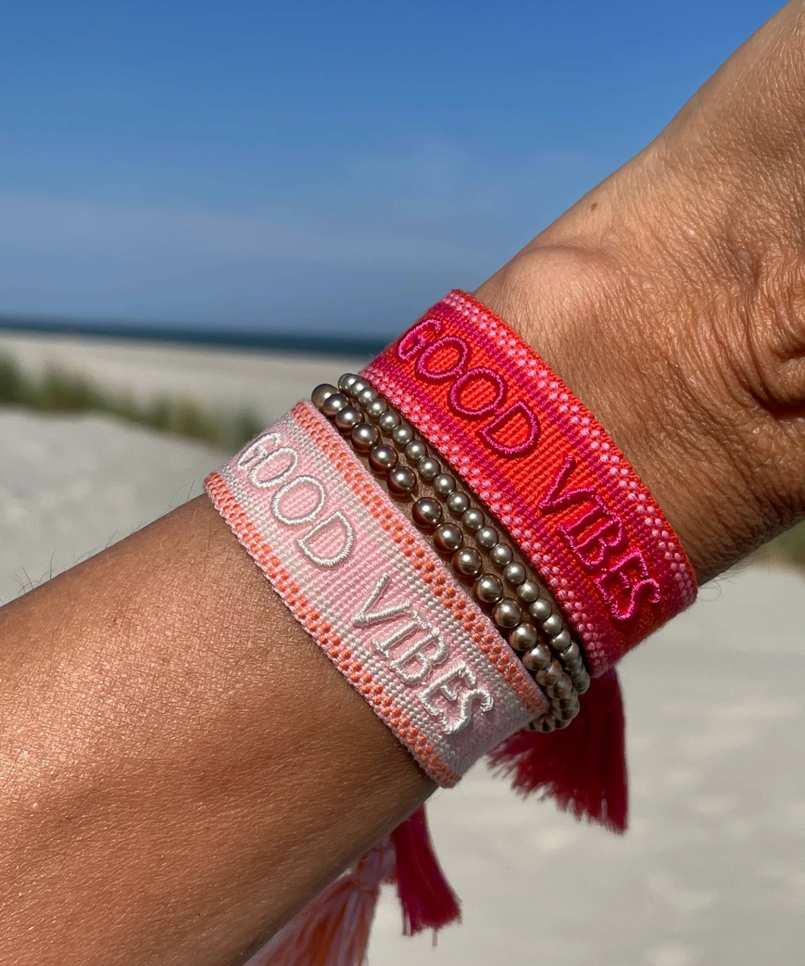 Close-up of adjustable rosy peach woven bracelet with embroidered “GOOD VIBES” lettering and sliding knot tassel cords.