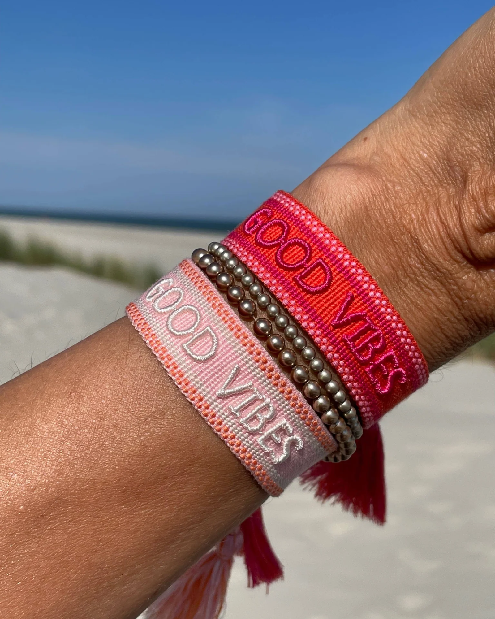 Close-up of adjustable rosy peach woven bracelet with embroidered “GOOD VIBES” lettering and sliding knot tassel cords.