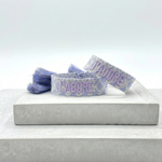 Close-up of adjustable purple and cream jacquard woven friendship bracelet with lilac embroidered “J’ADORE” lettering and sliding knot tassel cords.
