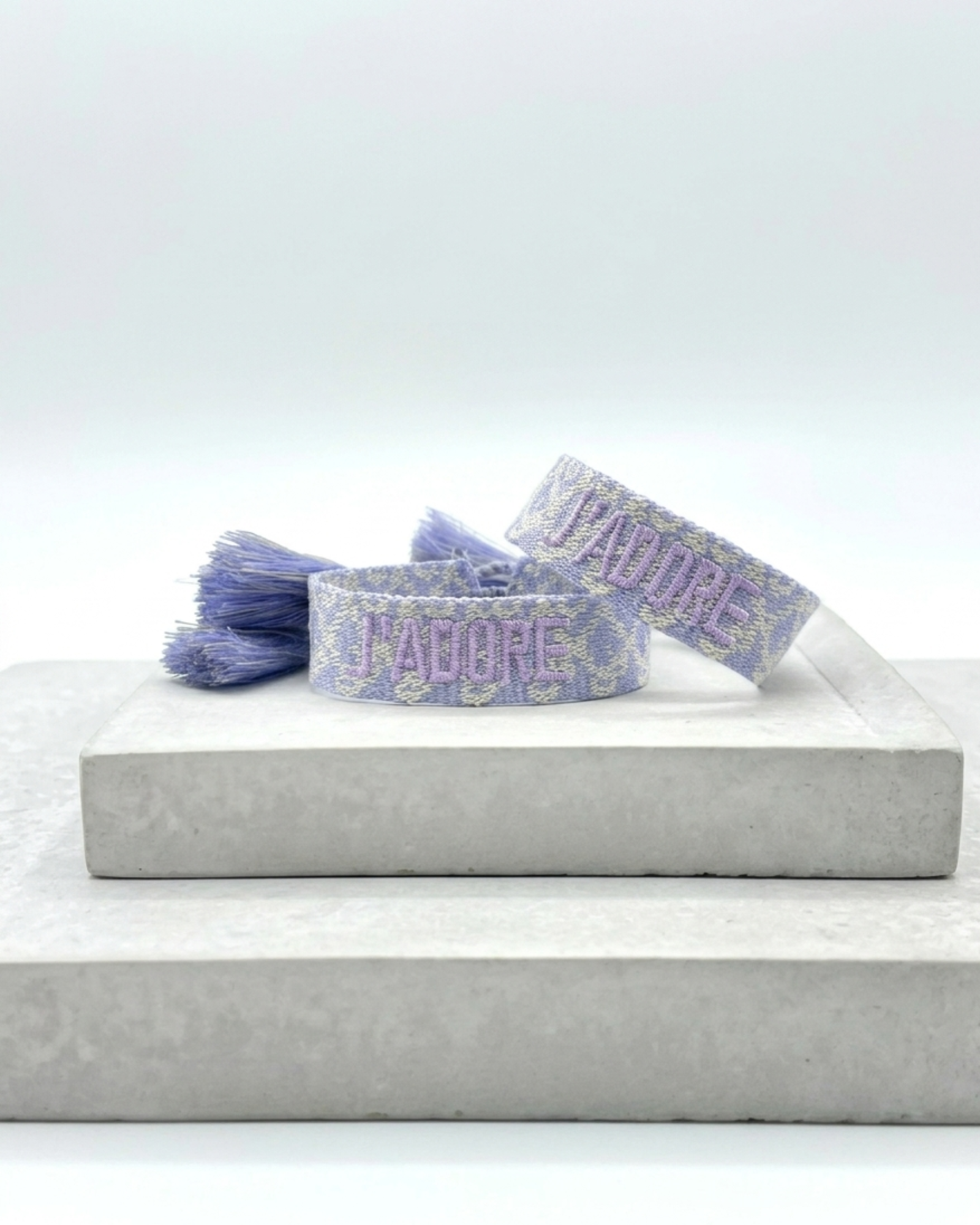 Close-up of adjustable purple and cream jacquard woven friendship bracelet with lilac embroidered “J’ADORE” lettering and sliding knot tassel cords.