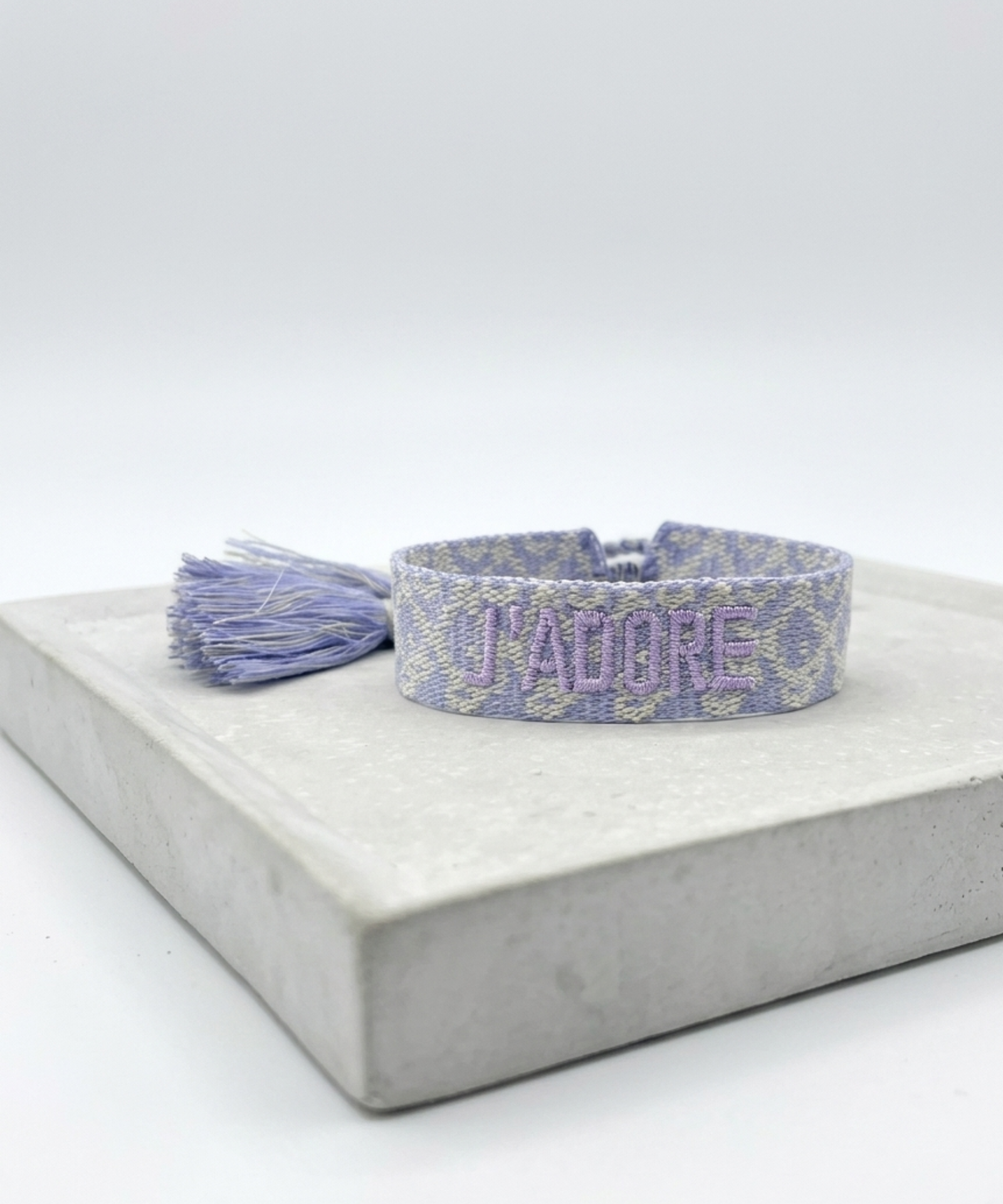 Close-up of adjustable lilac jacquard woven bracelet with embroidered “J'ADORE” lettering and sliding knot tassel cords.