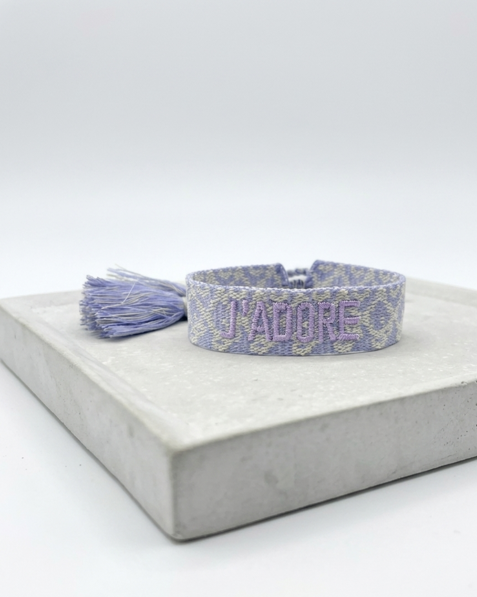 Close-up of adjustable lilac jacquard woven bracelet with embroidered “J'ADORE” lettering and sliding knot tassel cords.