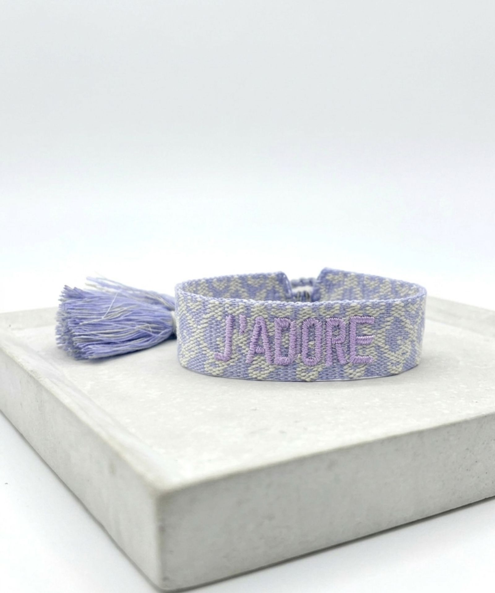 Close-up of adjustable purple and cream jacquard woven friendship bracelet with lilac embroidered “J’ADORE” lettering and sliding knot tassel cords.