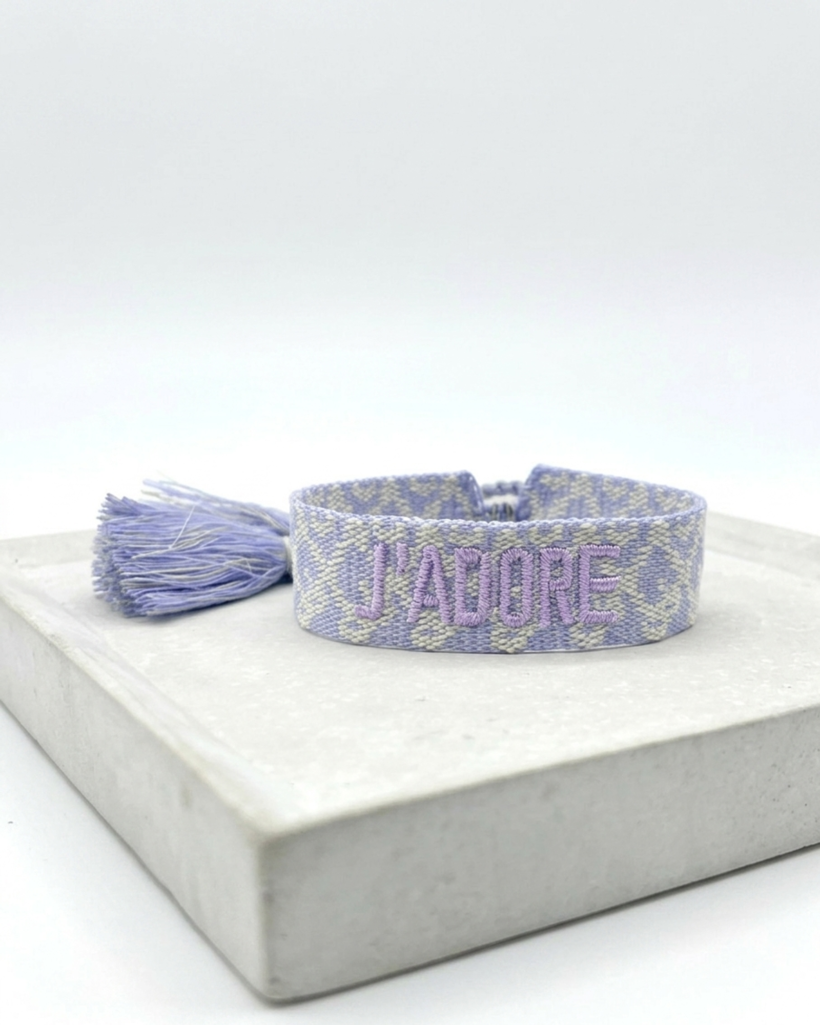 Close-up of adjustable purple and cream jacquard woven friendship bracelet with lilac embroidered “J’ADORE” lettering and sliding knot tassel cords.