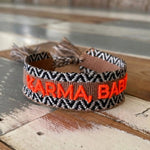 Close-up of adjustable woven boho friendship bracelet with neon orange “Karma, Baby!” embroidery and sliding tassel cords on a brown woven band.