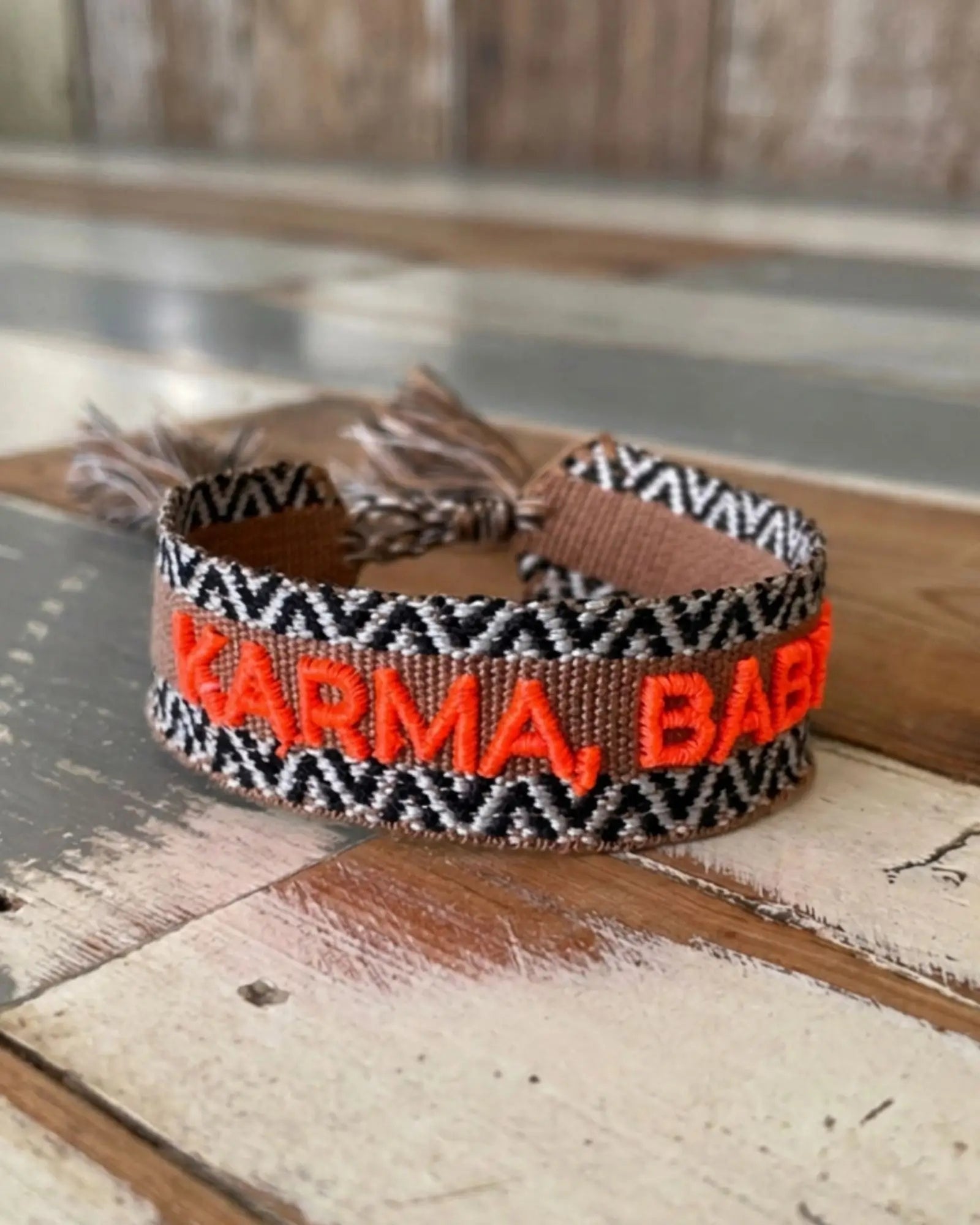 Close-up of adjustable woven boho friendship bracelet with neon orange “Karma, Baby!” embroidery and sliding tassel cords on a brown woven band.