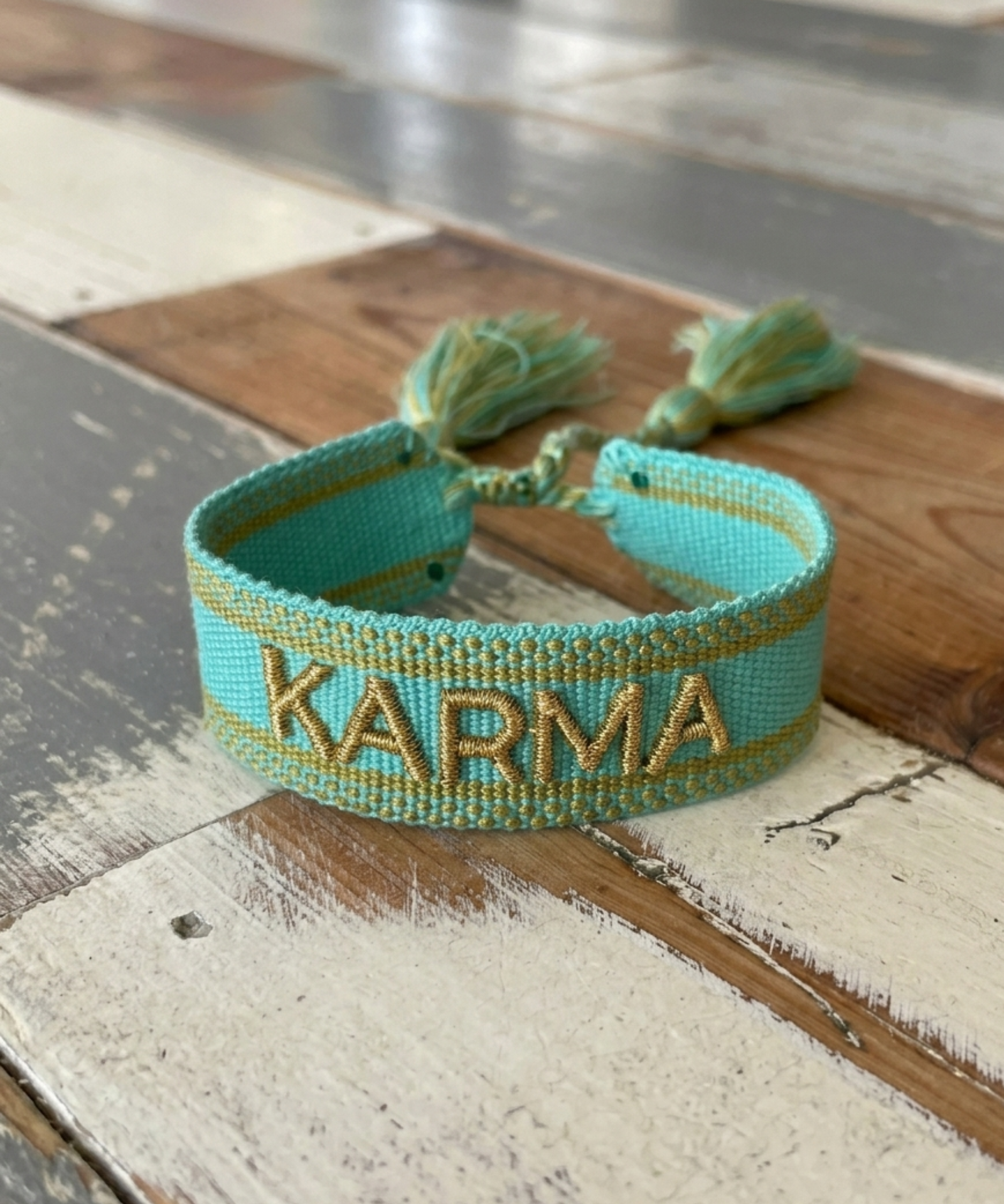 Close-up of adjustable turquoise woven bracelet with gold embroidered “KARMA” lettering and sliding knot tassel cords.