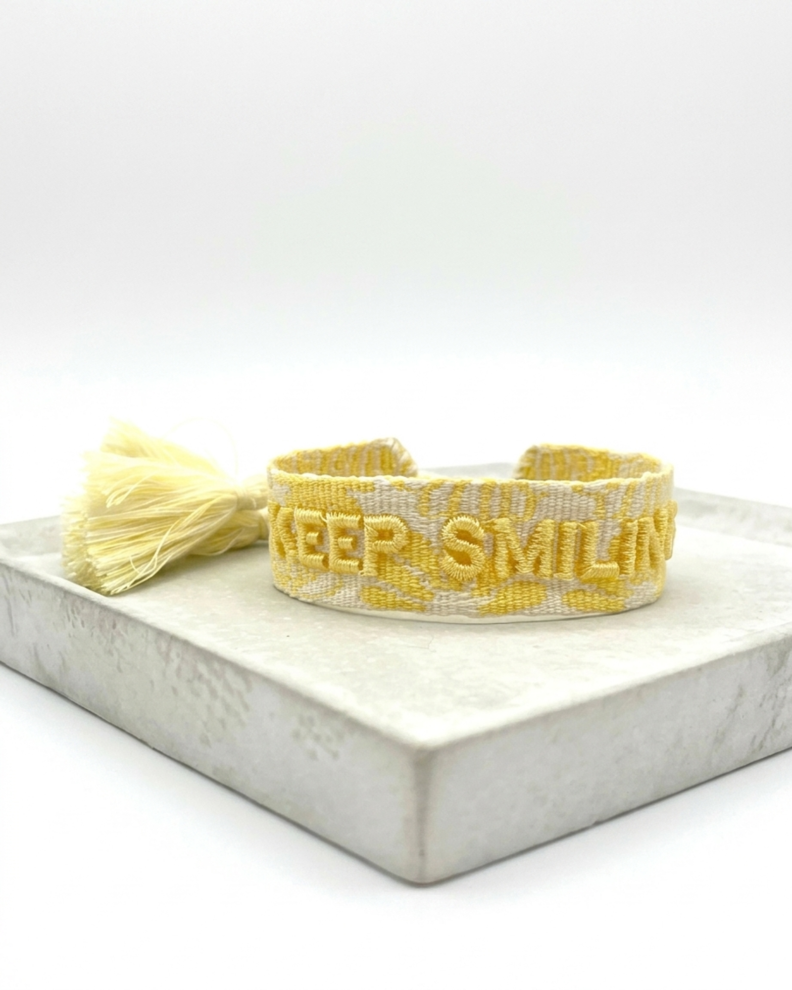 Close-up of adjustable yellow sorbet woven bracelet with embroidered “KEEP SMILING” lettering and sliding knot tassel cords.