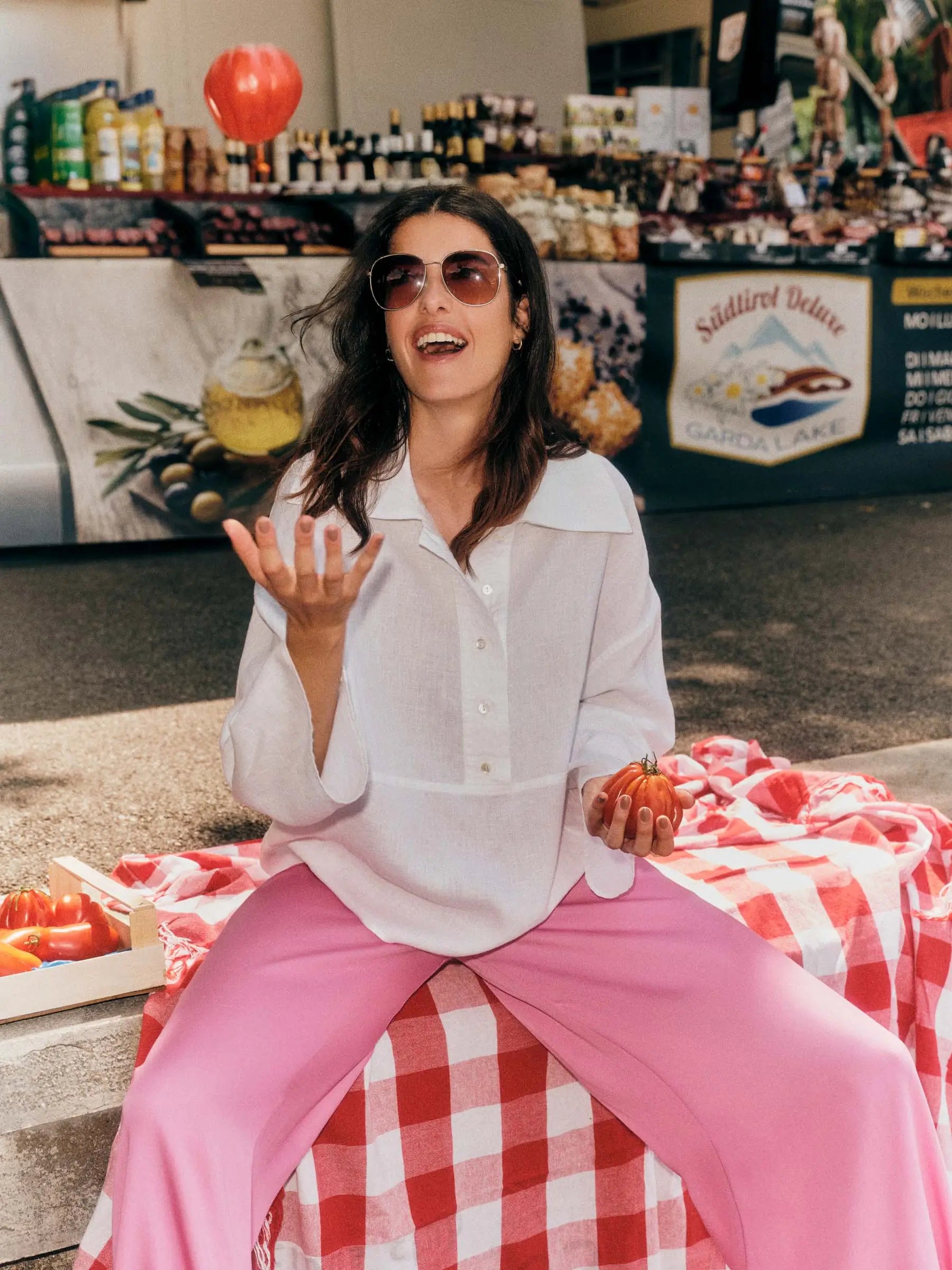 Woman in white shirt and pink pants by sustainable fashion brand Lanius sitting on a checkered blanket with produce at a market in Itay.