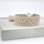 Close-up of adjustable light taupe woven bracelet with ecru embroidered “L'AMOUR” lettering, matching border, and sliding knot tassel cords.