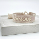 Close-up of adjustable light taupe woven bracelet with ecru embroidered “L'AMOUR” lettering, matching border, and sliding knot tassel cords.