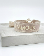 Close-up of adjustable light taupe woven bracelet with ecru embroidered “L'AMOUR” lettering, matching border, and sliding knot tassel cords.