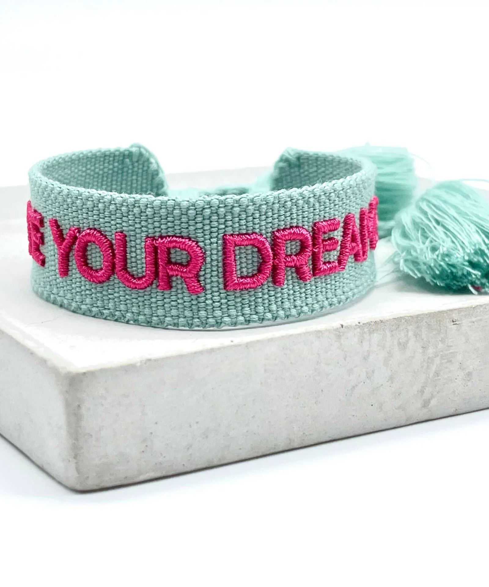 Close-up of adjustable sage green woven friendship bracelet with dark pink embroidered "LIVE YOUR DREAMS" lettering and sliding knot cords.