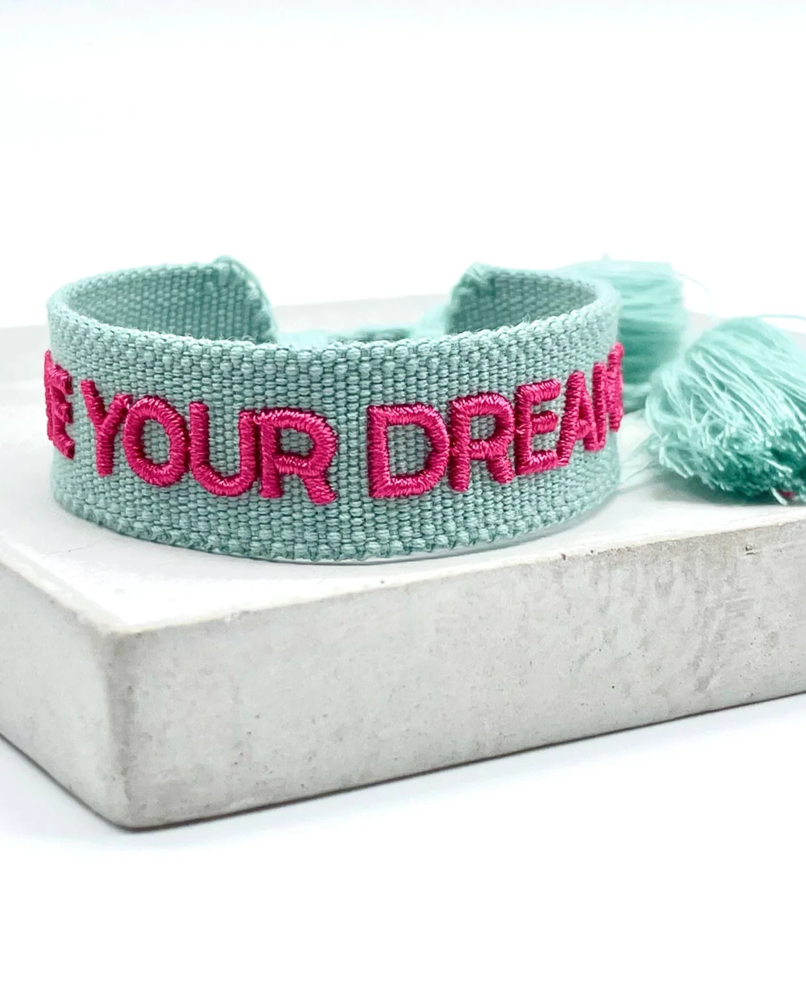 Close-up of adjustable sage green woven friendship bracelet with dark pink embroidered "LIVE YOUR DREAMS" lettering and sliding knot cords.
