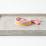 Close-up of adjustable salmon woven boho bracelet with offwhite border and orange embroidered “MORE VEUVE PLEASE” lettering, featuring sliding knot tassel cords.