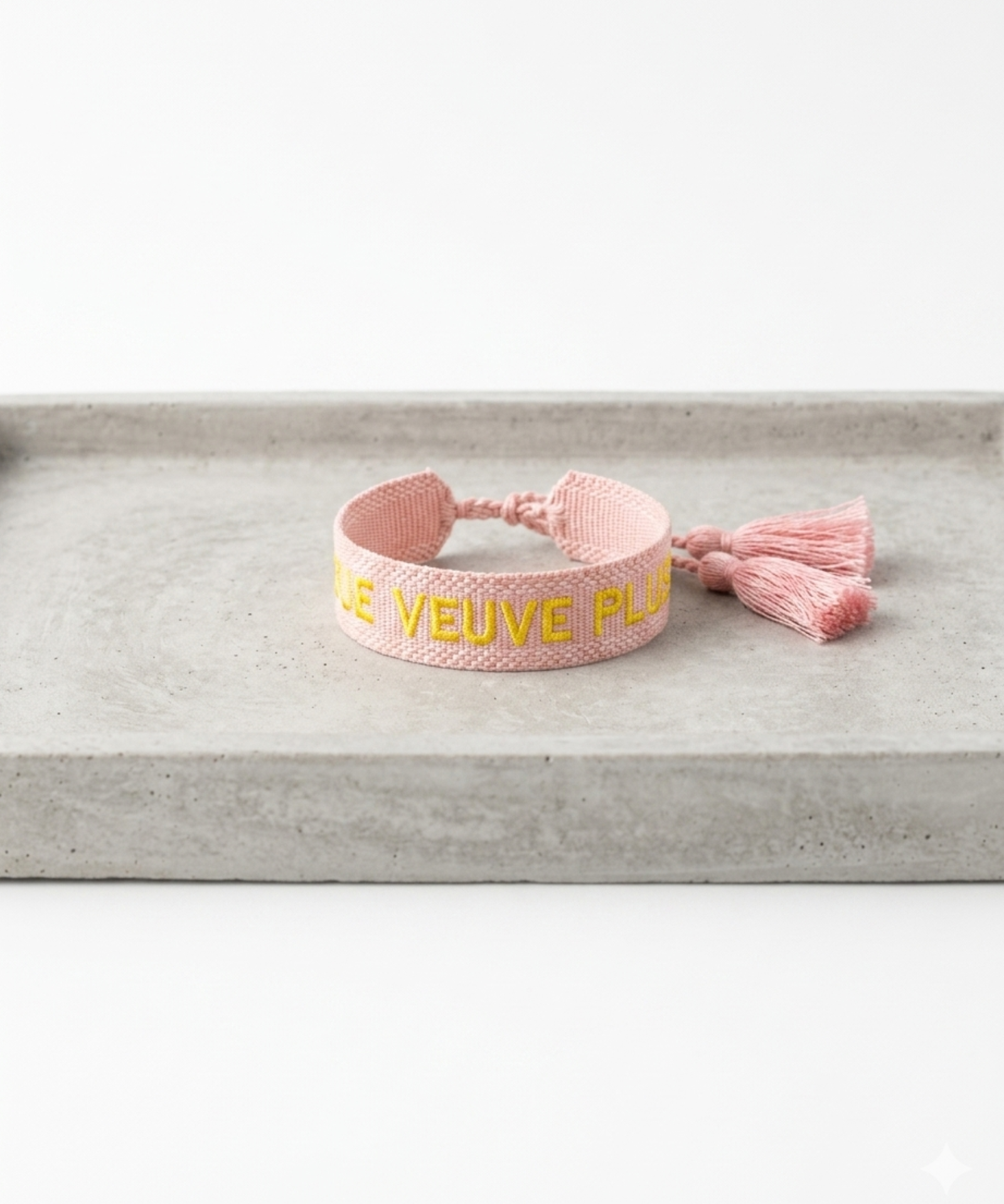 Close-up of adjustable salmon woven boho bracelet with offwhite border and orange embroidered “MORE VEUVE PLEASE” lettering, featuring sliding knot tassel cords.