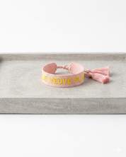 Close-up of adjustable salmon woven boho bracelet with offwhite border and orange embroidered “MORE VEUVE PLEASE” lettering, featuring sliding knot tassel cords.