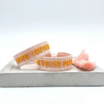 Close-up of adjustable salmon woven boho bracelet with offwhite border and orange embroidered “MORE VEUVE PLEASE” lettering, featuring sliding knot tassel cords.