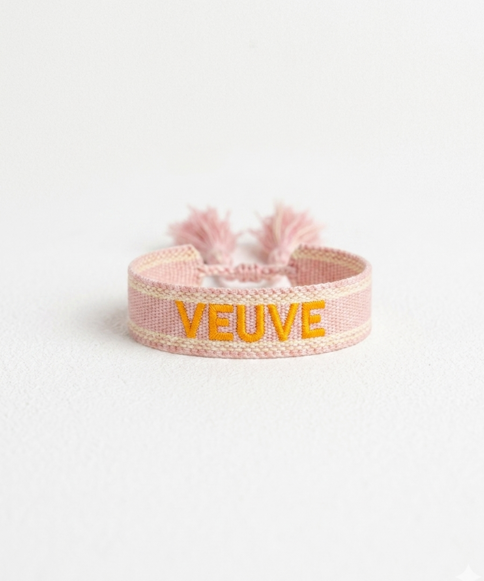 Close-up of adjustable salmon woven boho bracelet with offwhite border and orange embroidered “MORE VEUVE PLEASE” lettering, featuring sliding knot tassel cords.