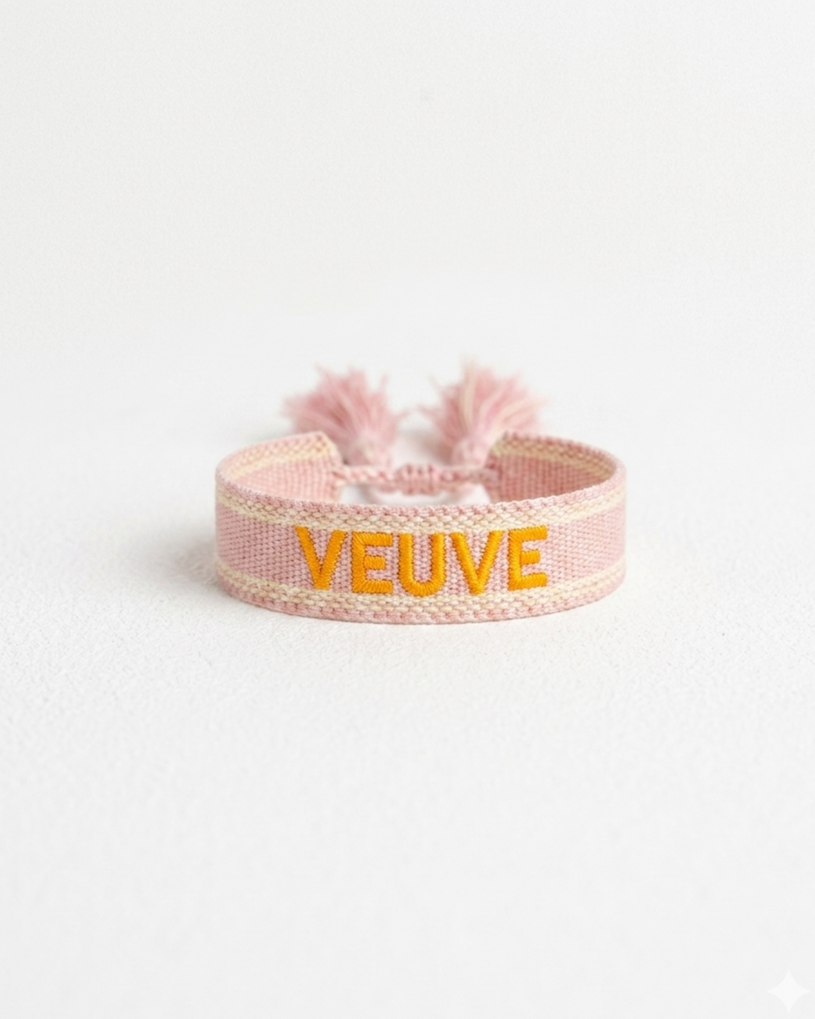 Close-up of adjustable salmon woven boho bracelet with offwhite border and orange embroidered “MORE VEUVE PLEASE” lettering, featuring sliding knot tassel cords.