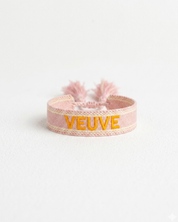 Close-up of adjustable salmon woven boho bracelet with offwhite border and orange embroidered “MORE VEUVE PLEASE” lettering, featuring sliding knot tassel cords.