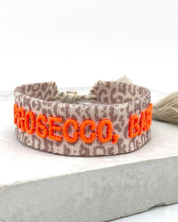Close-up of adjustable woven friendship bracelet with Leo pattern and neon orange embroidered "PROSECCO, BABY!" lettering with sliding knot cords.