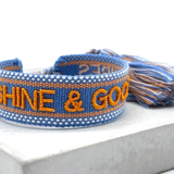 Close-up of adjustable blue and orange woven friendship bracelet featuring embroidered "SUNSHINE & GOOD VIBES" lettering and sliding knot tassel cords.